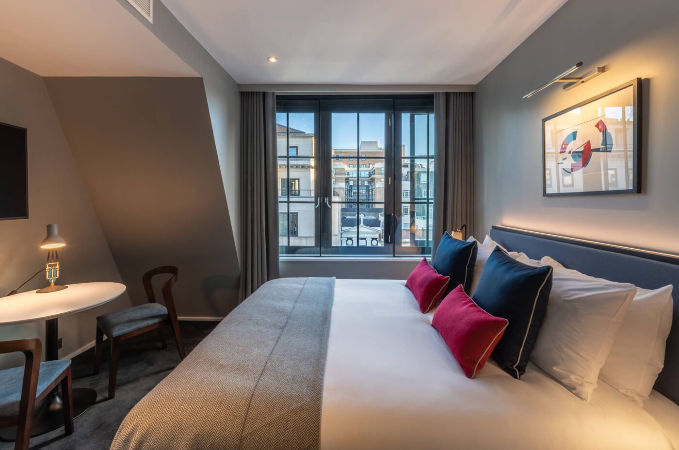 A modern hotel room with a large bed featuring white linens and colorful pillows, a small table with two chairs, a window with city views, and a framed abstract artwork on the gray wall.
