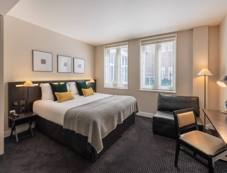 A modern, Central London hotel room at The Resident Soho with a large bed featuring grey bedding, white pillows, and yellow accent pillows. The room includes two windows, wall art above the bed, a black armchair, a desk with a chair, and sleek, ambient lighting. The carpet is dark grey.