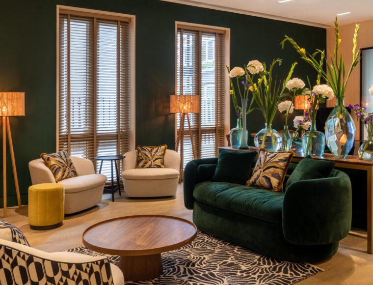 A modern living room with dark green walls and large windows at 4 star Soho hotel. The room features a green velvet sofa, beige chairs, a wooden coffee table, and a zebra-patterned rug. Vases of tall flowers sit on a console table while wooden floor lamps with decorative shades illuminate the space, reminiscent of The Resident Soho's chic decor.