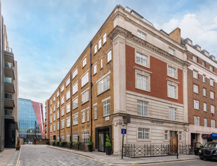  A view of a street corner in the city, blending modern and historic architecture. A red-brick building with white trim occupies the corner. This is The Resident's Victoria hotel in London. Cobblestones pave the street, while modern buildings line it. A glass-fronted structure stands in the background.