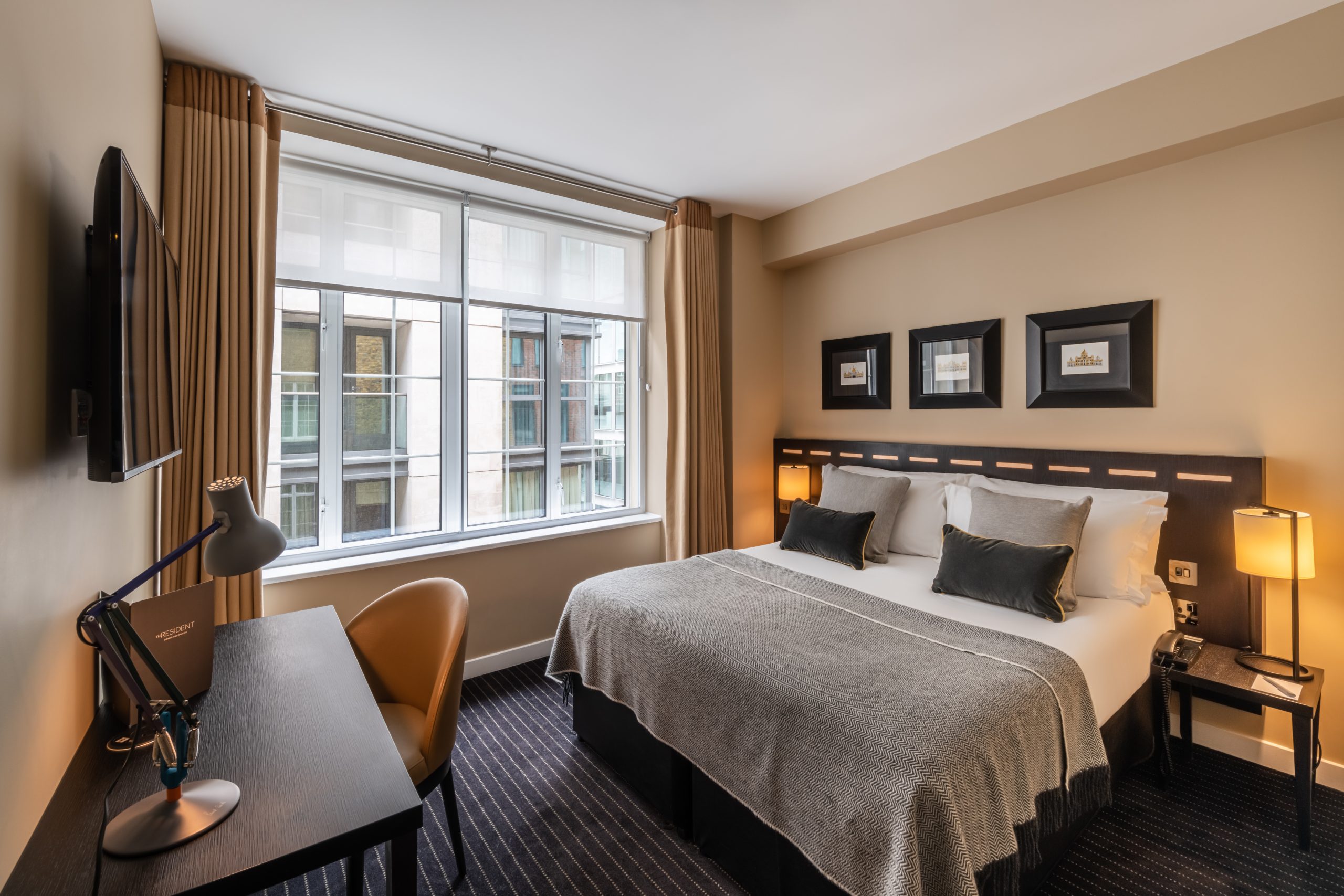 A cozy hotel room at The Resident Victoria features beige walls, a large bed adorned with white and grey linens, and several pillows. The room includes a work desk with a chair and desk lamp, a wall-mounted TV, and two bedside tables with lamps. A large window allows natural light to brighten up the space.