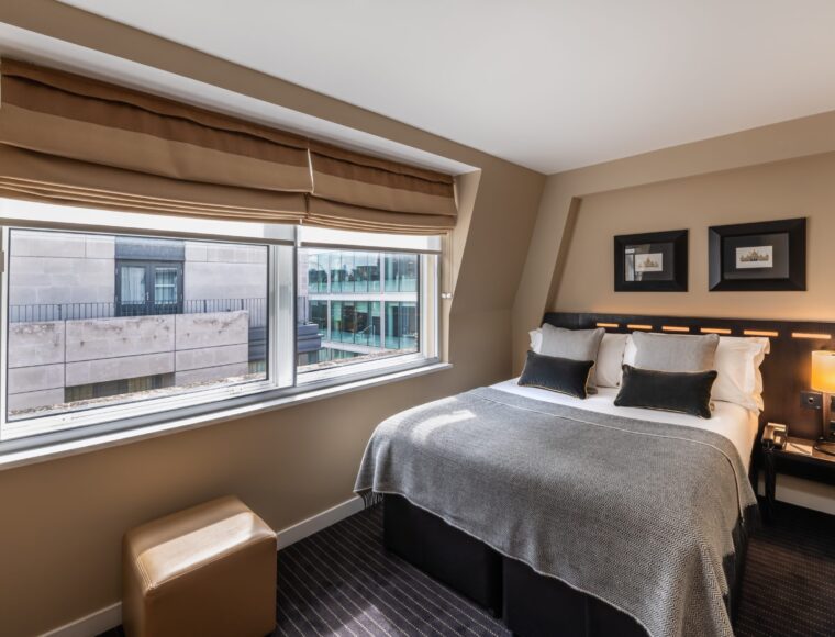 A cozy hotel room at The Resident Victoria with a double bed adorned with grey and white bedding and black pillows. The room features two windows with beige blinds, framed wall art, a bedside table with a lamp, and a small brown ottoman by the window. The exterior view shows modern buildings.