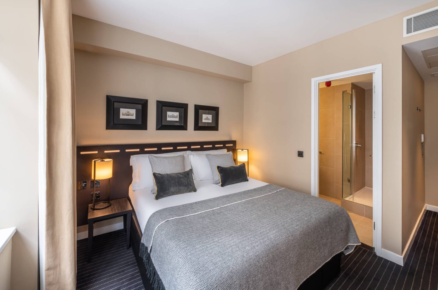 A neatly made bed with grey bedding and four pillows is situated in a modern hotel room at The Resident Victoria. This London hotel room has beige walls, two bedside lamps, a dark wooden headboard, and three framed pictures above the bed. A door on the right leads to a bathroom with a glass shower.