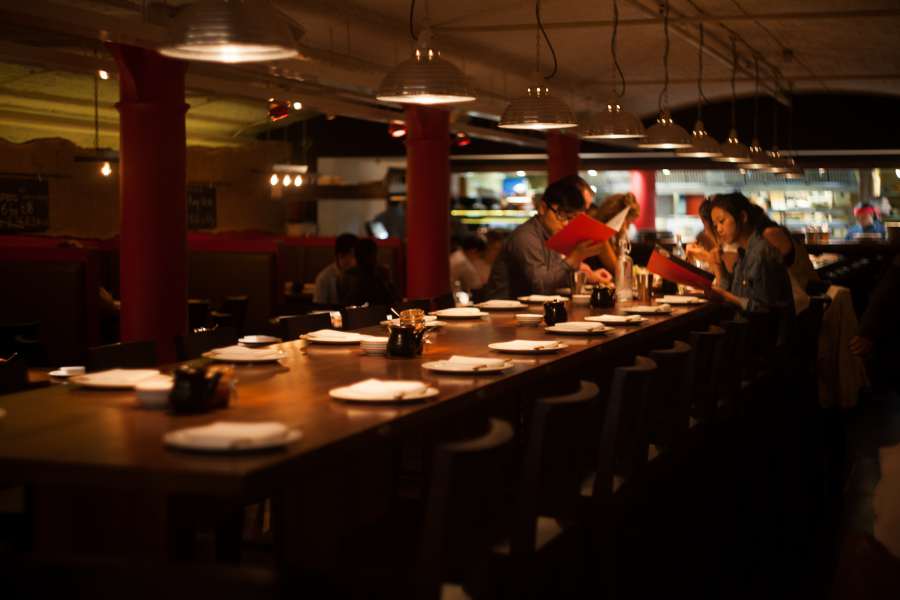 Dimly lit restaurant with a long wooden table set with white plates and utensils. Red columns line the room, and various people are engaged in conversation near the bar area, which is illuminated. The ambient lighting creates a warm and cozy atmosphere. The restaurant is nearby The Resident Covent Garden.