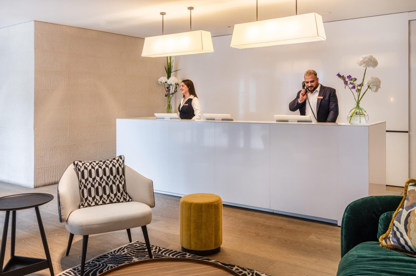 A modern hotel lobby at The Resident Covent Garden features a front desk with two staff members behind it; a woman and a man, both using phones. The area has stylish furniture, including a white armchair with a patterned cushion, a yellow pouf, and a green sofa. Soft lighting creates a welcoming atmosphere.