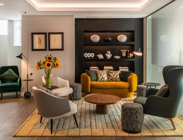 A modern living room with a mustard sofa, two gray chairs, and a green armchair. A wooden coffee table sits on a striped rug. Sunflowers are in a vase, and a shelf with decorative items is in the background. A floor lamp and artwork are on the left wall.