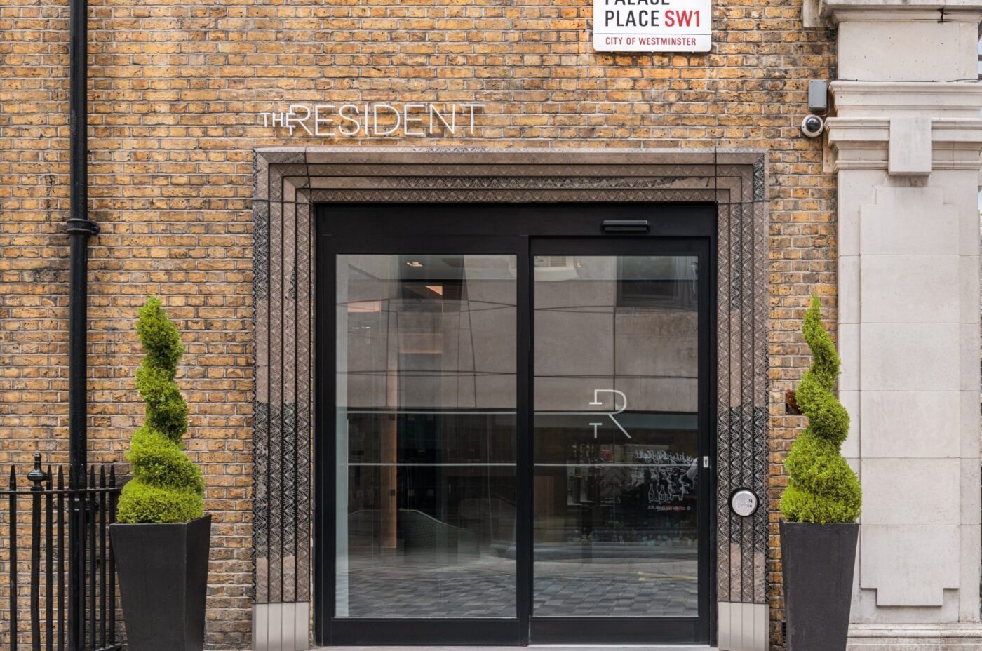 Glass entrance door of The Resident hotel with two spiral-shaped potted topiary plants on either side, set in a brick building. A street sign above reads “Palace Place SW1, City of Westminster.”.