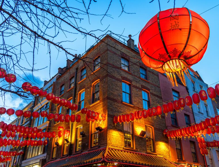 A city street is adorned with numerous red lanterns hanging across the sky. A large red lantern takes prominence in the foreground. The brick building below is warmly lit, set against a twilight sky, creating a festive ambiance.