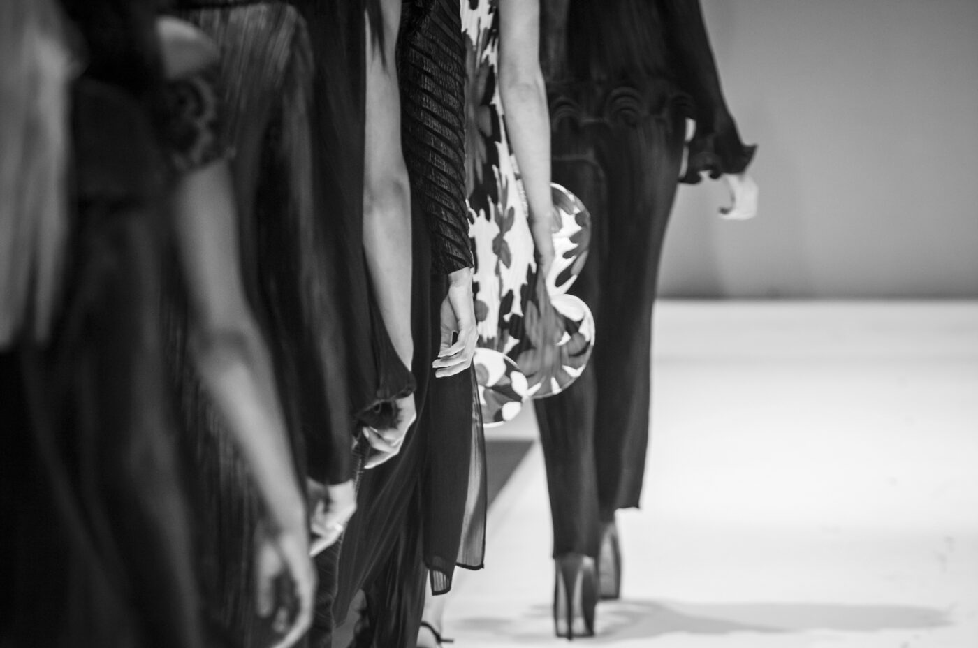 A black-and-white photo of models walking down a runway. The focus is on their legs and clothing, showcasing various textures and patterns. The models wear high heels and elegant outfits, creating a sleek and stylish scene.