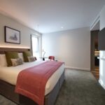 A modern hotel room featuring a large bed with white linens, green and white pillows, and a red blanket. A wooden headboard adorns the bed. There's a desk with a lamp, a chair, and a wall-mounted art piece. The room has soft lighting and carpeted flooring.