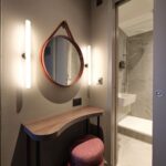A modern bathroom entrance featuring a black countertop with a wavy surface, a red cushioned stool, and a round mirror with a leather strap. The interior bathroom has gray tiles, a white sink, and a shower area with a glass door.
