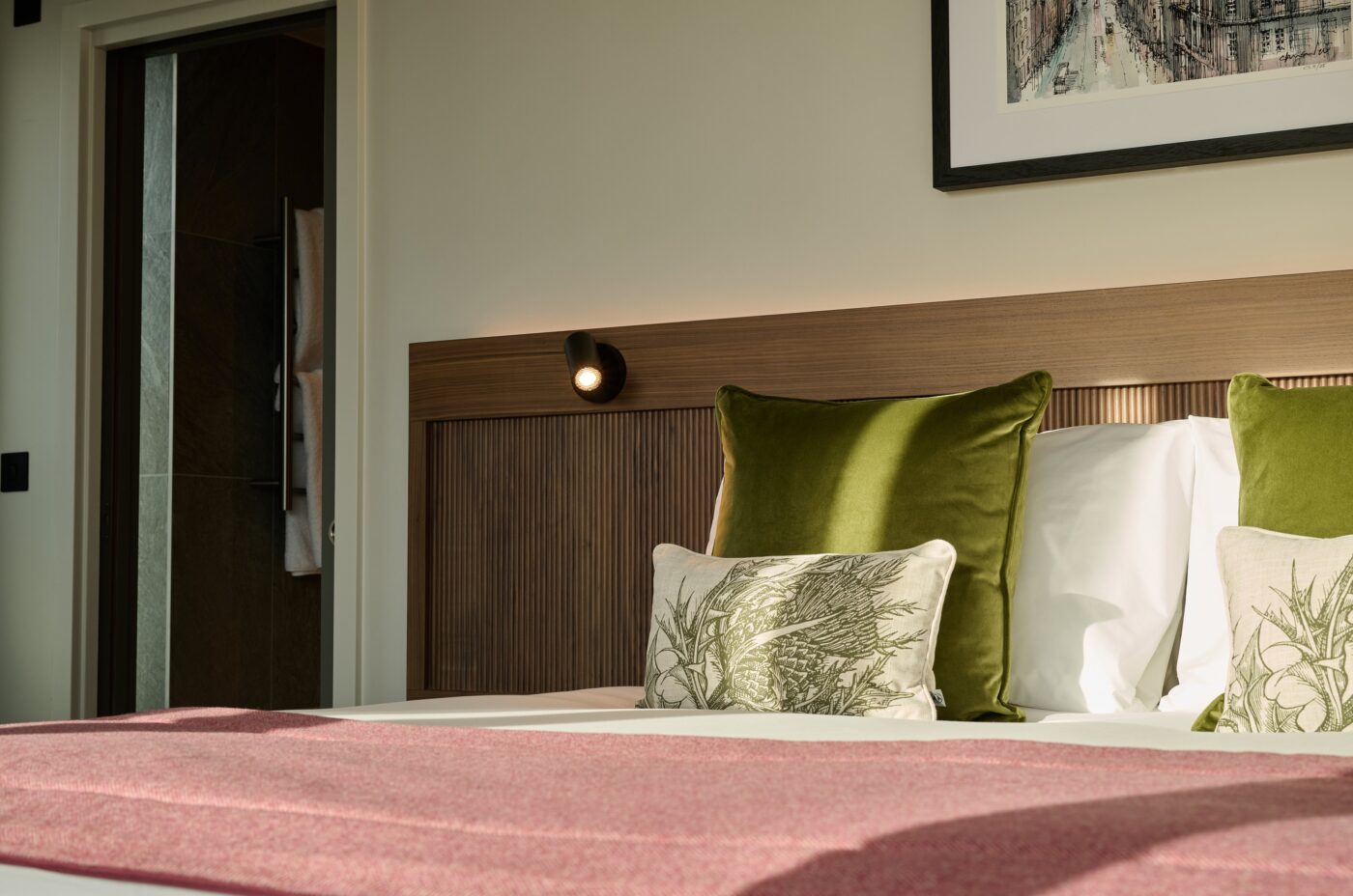 A cozy bedroom features a neatly made bed with green and patterned pillows, a pink blanket, and a wooden headboard. A framed picture hangs on the wall, and sunlight streams in from a nearby window, creating a warm atmosphere.