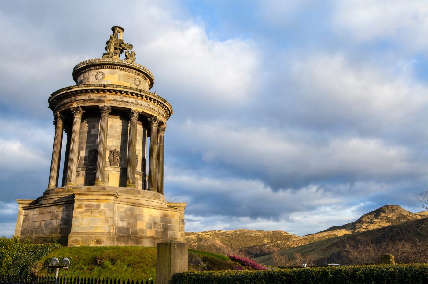 A neoclassical monument with columns stands prominently on a grassy hill, set against a backdrop of partly cloudy skies and rolling hills. As if preparing for Burns Night, warm sunlight highlights the structure, enhancing its intricate architectural details.