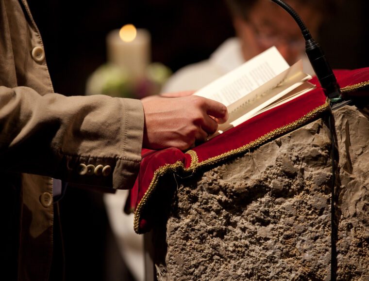 A person in a tan coat stands at a stone lectern with a red velvet cover, reading from an open book. A lit candle and another person blurred in the background suggest a formal setting, reminiscent of Burns Night celebrations.