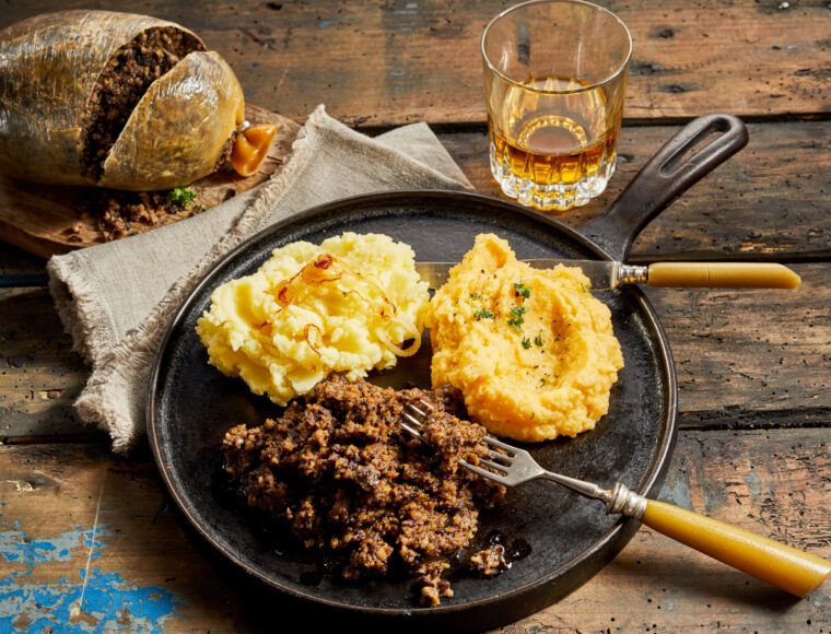 A rustic wooden table showcases a dish of haggis, mashed potatoes, and turnips on a metal plate with cutlery. A glass of whiskey and a whole haggis on a cloth complete the scene, perfectly capturing the essence of Burns Night in this traditional Scottish meal setting.