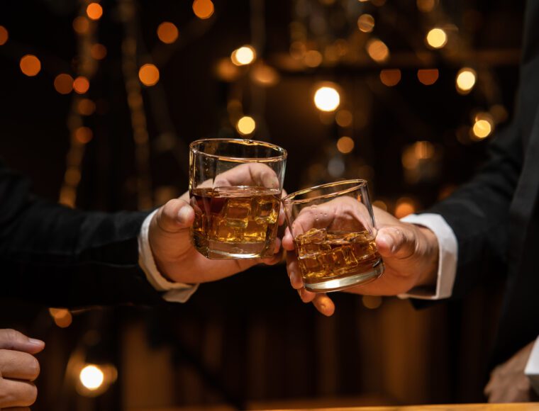 Two people clink glasses of whiskey in a dimly lit setting reminiscent of Burns Night, with warm, blurred lights in the background. One is dressed in a black suit, the other in a dark jacket. A hand rests thoughtfully on the table nearby.