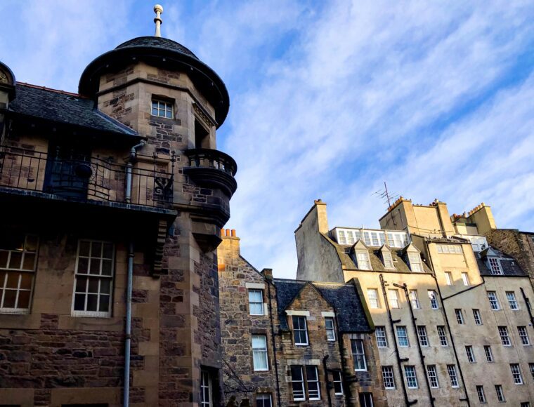 A view of historic stone buildings with varied architectural styles under a blue sky evokes the spirit of Burns Night. The foreground features a round tower with a balcony, while the background showcases multi-story structures adorned with chimneys and a mix of windows.