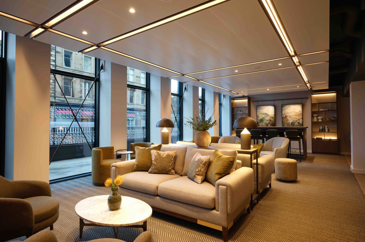 Modern lounge area with comfortable sofas and armchairs, decorated with cushions and plants. Large windows provide natural light, and a small round table with flowers is centered. A bar area with stools and artwork is visible in the background.
