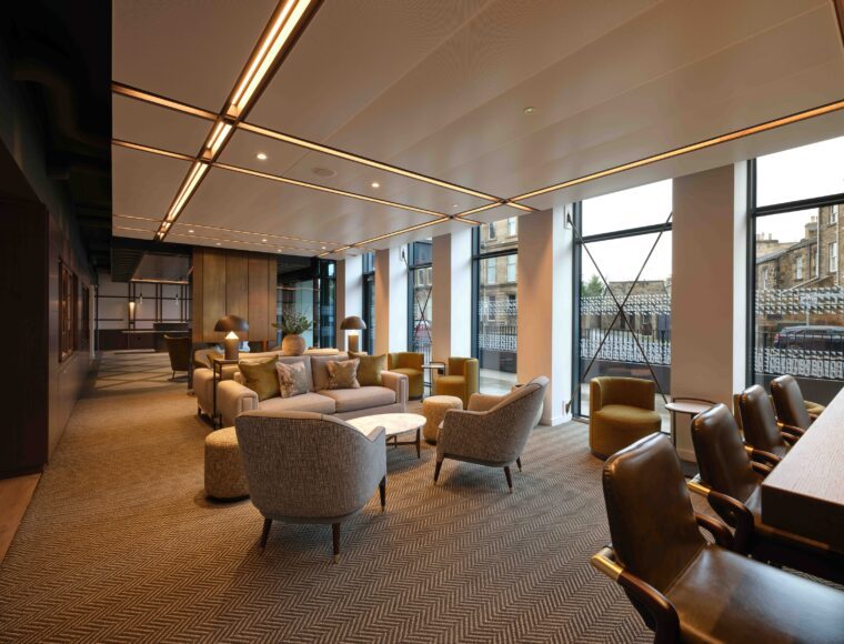 Modern lounge with large windows, contemporary furniture, and neutral tones. The seating area includes armchairs, sofas, and a coffee table atop a patterned carpet. Tall windows allow natural light, and a view of an urban landscape is visible outside.