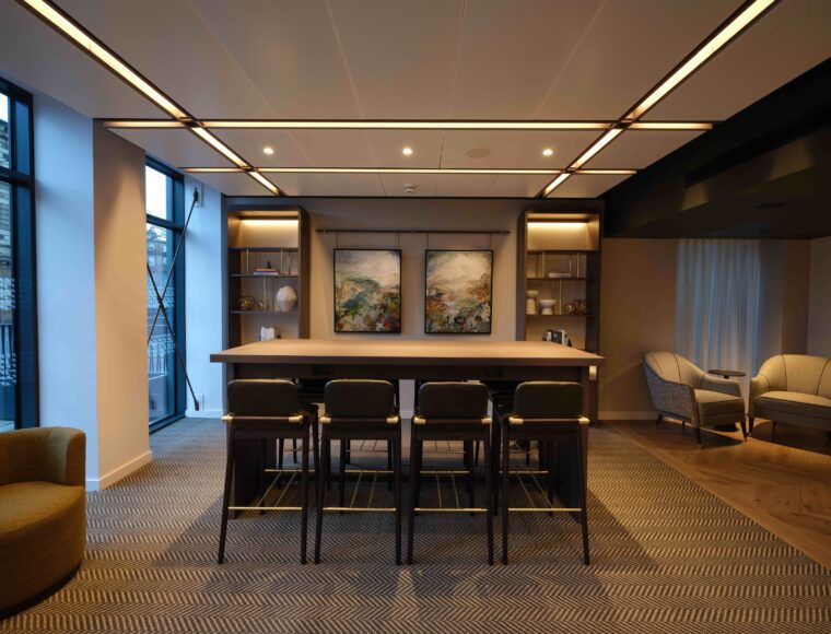 A modern office lounge with a long table surrounded by chairs, contemporary art on the wall, shelves with decorative items, and two armchairs in the corner. Large windows provide natural light, and recessed lighting adds a warm ambiance.