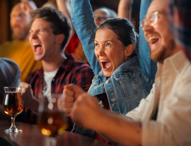 A group of excited people sit at a bar, cheering and celebrating. Beverages are on the table, and everyone is focused on something off-camera with joyful expressions.