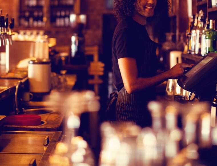 A person with curly hair is smiling while using a register behind a bar. The bar is lined with various bottles and glasses, and the lighting is warm and dim, creating a cozy atmosphere.