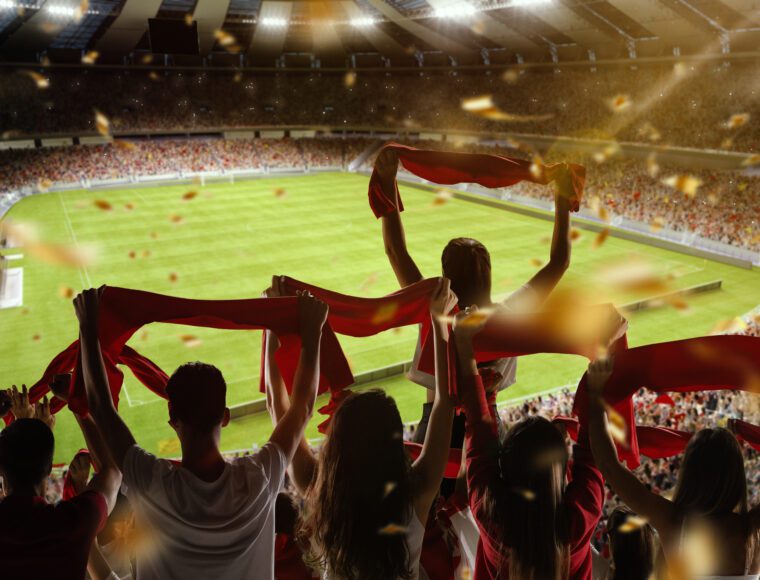 A group of enthusiastic fans with red scarves is cheering on Liverpool FC in the stands of a , Anfield stadium. The well-lit field is visible in the background, with confetti falling, creating a festive atmosphere.