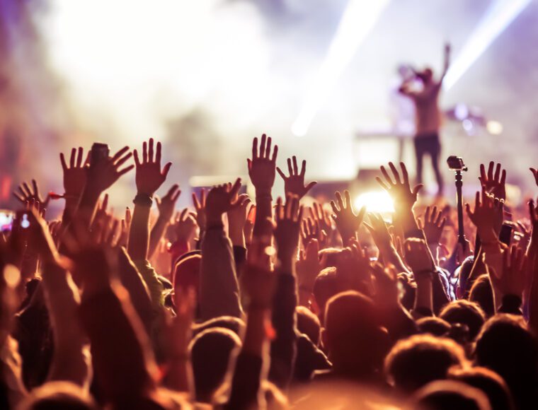 A lively crowd at a music festival concert with numerous hands raised in the air. A performer on stage is illuminated by bright lights, creating a vibrant and energetic atmosphere.