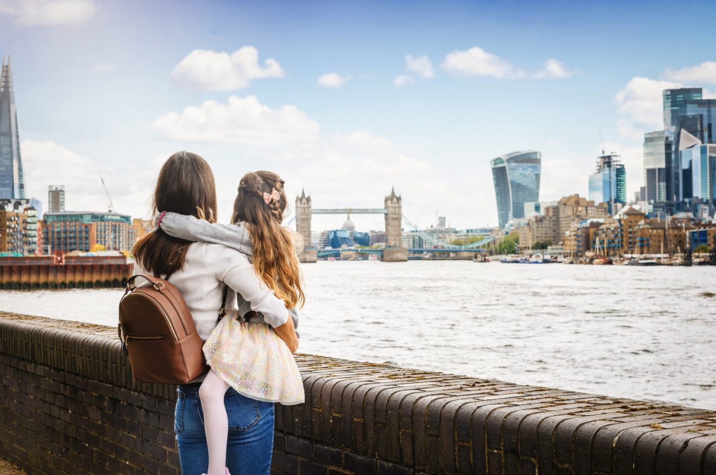 A woman with a brown backpack holds a young girl as they look across the Thames River at the Tower Bridge and London's modern skyline on a clear day.