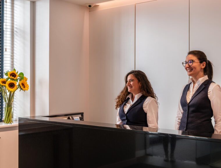 Two women in matching uniforms stand smiling behind a black reception desk in a modern office lobby. A vase of sunflowers sits on the windowsill to the left, and sunlight streams in through the window blinds.